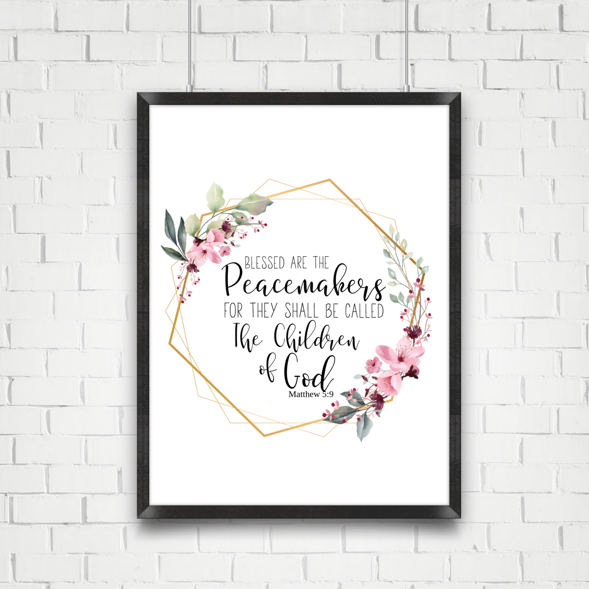 Black frame hanging on a white brick wall. Print in the frame displays a quote from Matthew 5:9 KJV that says Blessed are the peacemakers for they shall be called the Children of God. the quote is surrounded by a gold  geometric frame with pink flowers and greenery. 