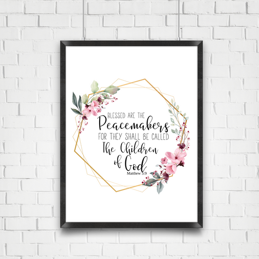Black frame hanging on a white brick wall. Print in the frame displays a quote from Matthew 5:9 KJV that says Blessed are the peacemakers for they shall be called the Children of God. the quote is surrounded by a gold  geometric frame with pink flowers and greenery. 