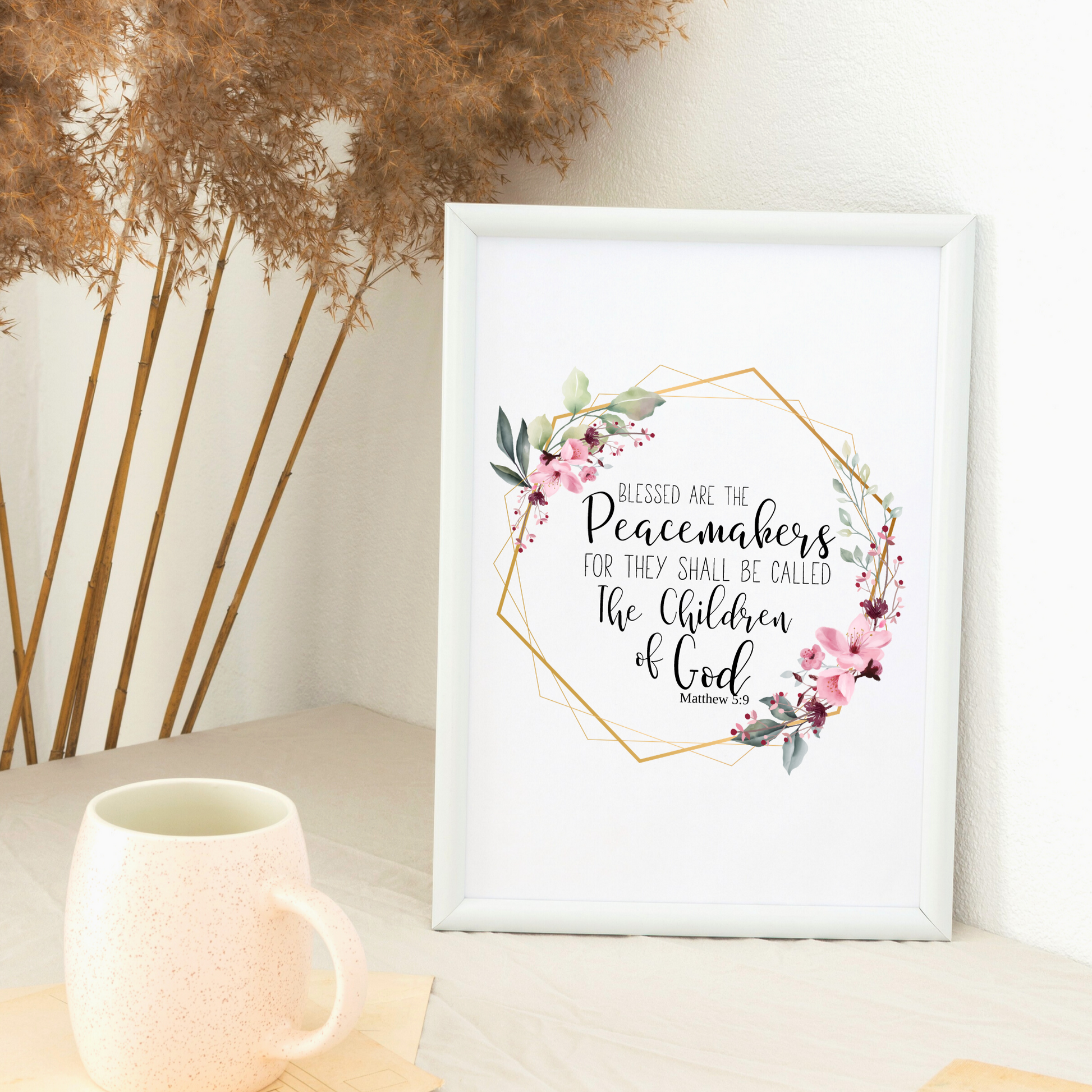 White frame sitting on a table with a white coffee cup sitting next to the frame and Pompas grass behind the table. Print in the frame displays a quote from Matthew 5:9 KJV that says Blessed are the peacemakers for they shall be called the Children of God. the quote is surrounded by a gold  geometric frame with pink flowers and greenery. 